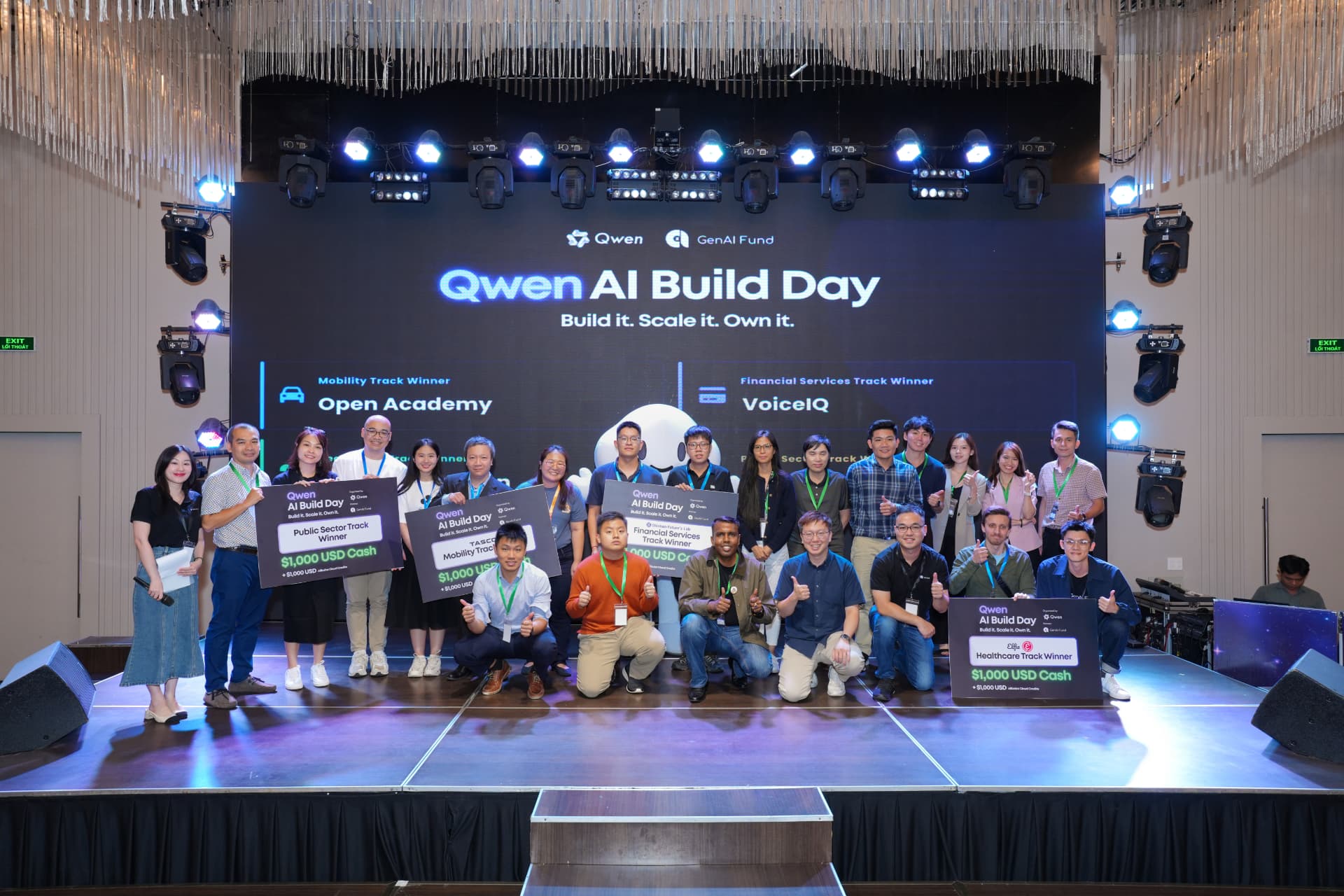 Qwen AI Build Day: track winners on stage holding prize boards for the Mobility, Financial Services, Public Sector and Healthcare tracks under the 'Build it. Scale it. Own it.' banner.