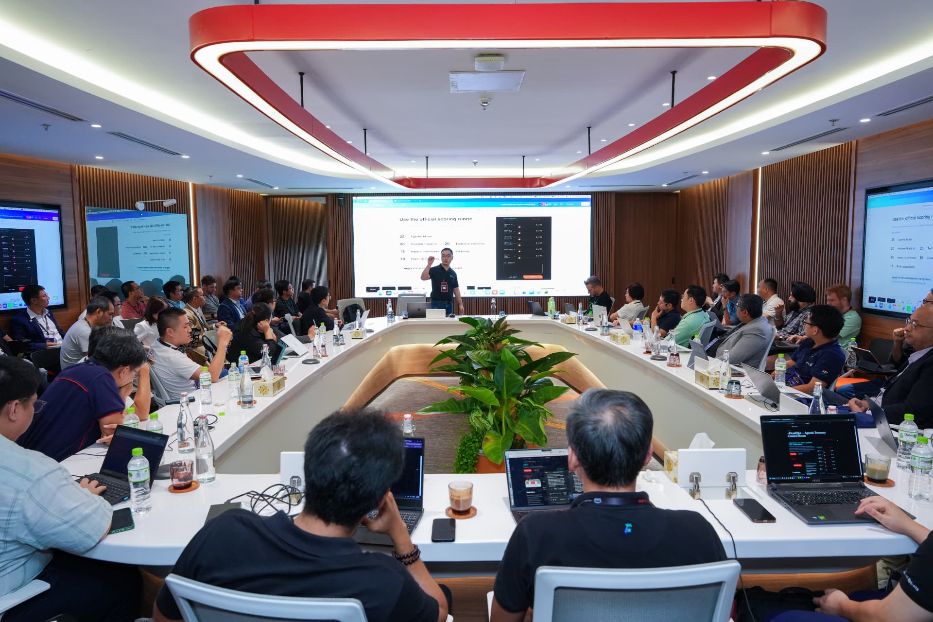 Enterprise teams filling a boardroom with laptops open while a facilitator presents the official scoring rubric during a GenAI Fund agentic AI build session.